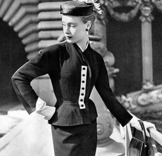 1952 Model in black wool suit, collarless jacket shows a hint of white at the neck and has a white piqué tab for the buttons, by Givenchy